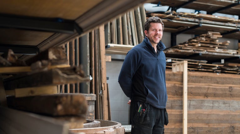Callum McCaffrey wears a black fleece and work trousers and stands in front of shelving full of wooden panelling